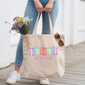 Op de afbeelding: Een beige canvas draagtas met een afbeelding van kleurrijke boeken op een rij en de tekst "well read." in roze letters. De tas heeft twee schouderbanden en wordt getoond met een bruine zonnebril die aan een van de banden hangt.