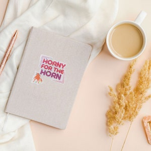 May include: A beige notebook with a sticker that says "Horny for the Horn" in a colorful, retro font. The sticker also features a cartoon unicorn.