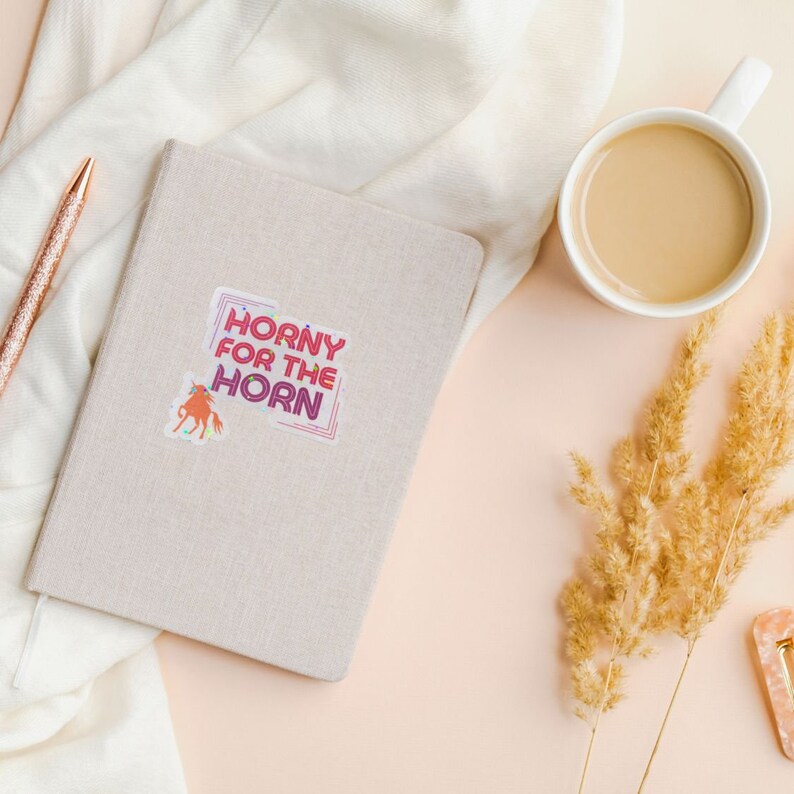 May include: A beige notebook with a sticker that says "Horny for the Horn" in a colorful, retro font. The sticker also features a cartoon unicorn.