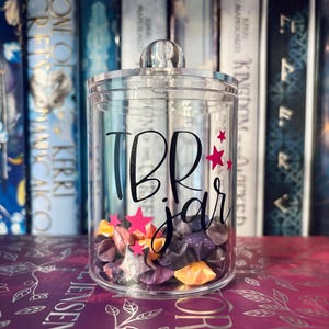 May include: A clear acrylic jar with a silver lid and filled with colourful origami stars. The jar is labelled "TBR jar" in black script with pink star accents. The jar is displayed in front of a bookshelf.