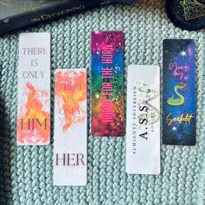 May include: Four colorful bookmarks with different designs and text. The bookmarks feature a snake, a crown, a fire design, and a floral design. The text on the bookmarks includes "There is only him", "There is only her", "Horny for the horn", and "Almighty Sovereign A.S.S Sogieity Snaketet".