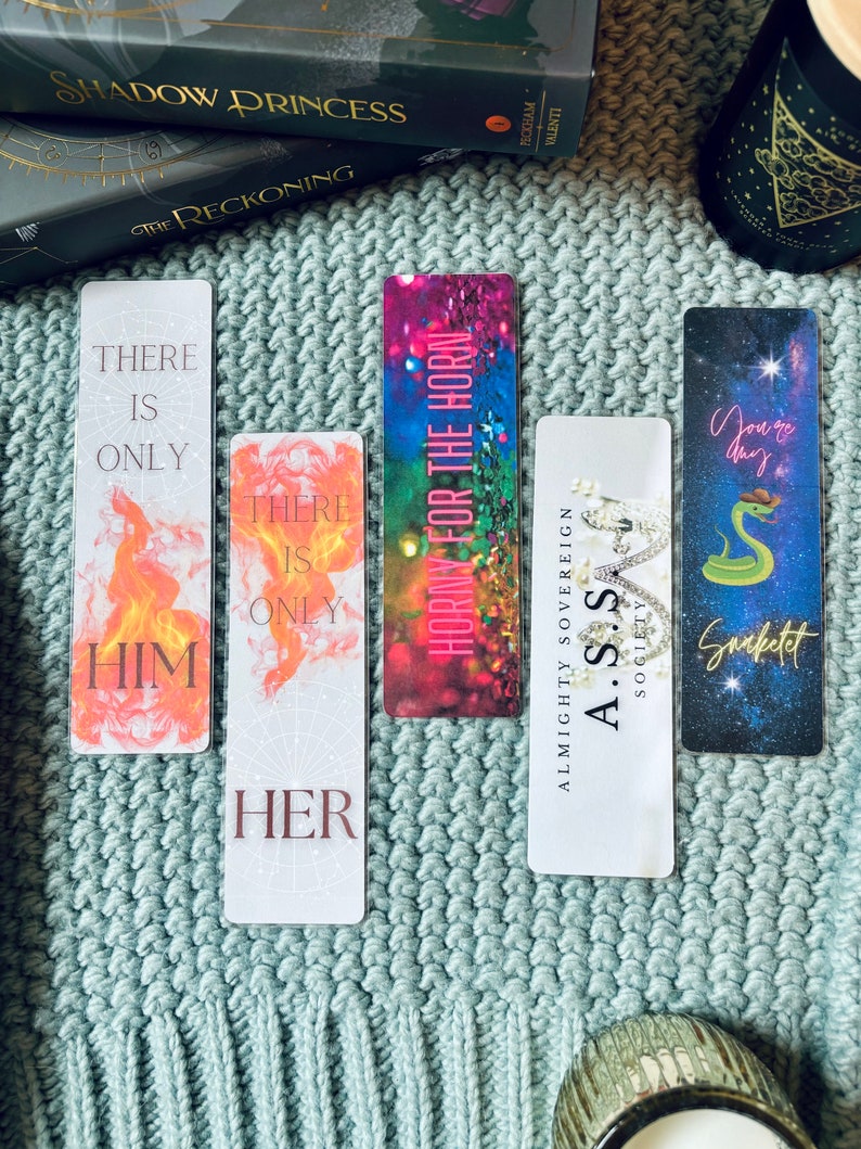May include: Four colorful bookmarks with different designs and text. The bookmarks feature a snake, a crown, a fire design, and a floral design. The text on the bookmarks includes "There is only him", "There is only her", "Horny for the horn", and "Almighty Sovereign A.S.S Sogieity Snaketet".