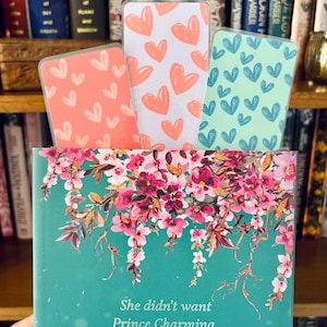 May include: Three heart-patterned bookmarks in coral, white, and teal, with a book featuring floral art and the text "She didn't want Prince Charming. She wanted the demon."