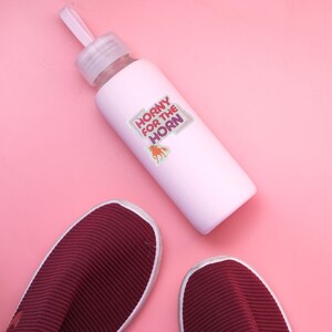 May include: A pink water bottle with a sticker that says "Horny for the Horn" with a cartoon unicorn on it. The bottle is on a pink surface next to two burgundy shoes.