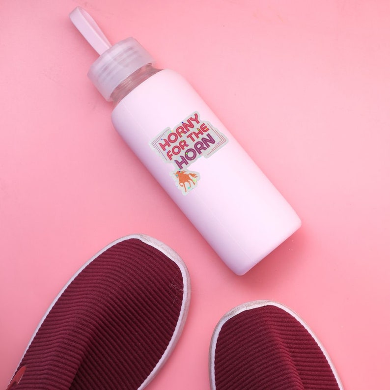May include: A pink water bottle with a sticker that says "Horny for the Horn" with a cartoon unicorn on it. The bottle is on a pink surface next to two burgundy shoes.