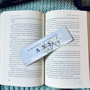 May include: A close-up of a bookmark with a white background and a silver border. The bookmark features a black and white illustration of a woman with a crown and the text "Almighty Sovereign A.S.S. Society".
