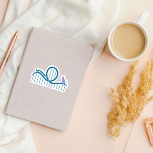 May include: A die-cut sticker featuring a blue roller coaster track design with a small purple cat silhouette sitting on the track. The sticker has a white border and is placed on a light grey notebook next to a pen and a cup of coffee.
