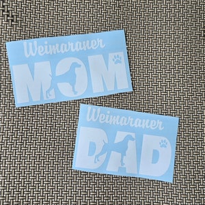 May include: Two white vinyl decals with the words "Weimaraner Mom" and "Weimaraner Dad" with a paw print and a silhouette of a Weimaraner dog.