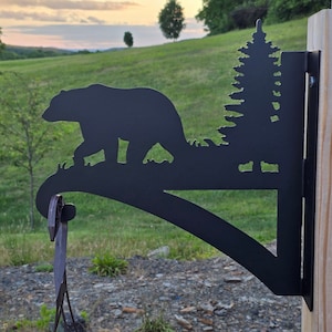 May include: Black metal silhouette of a bear walking through a forest with a pine tree in the background. The silhouette is attached to a curved metal bracket that is mounted to a wooden post.