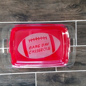 May include: A clear glass baking dish with red handles. The dish has a white football design with the text "GAME DAY CASSEROLE" etched into it.