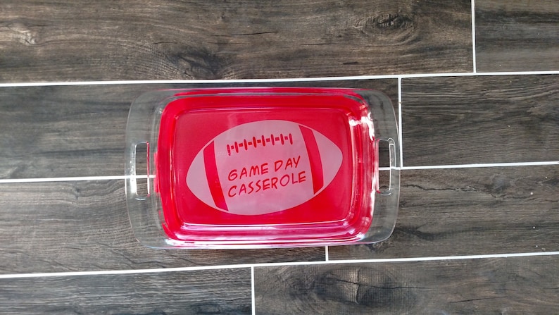 May include: A clear glass baking dish with red handles. The dish has a white football design with the text "GAME DAY CASSEROLE" etched into it.
