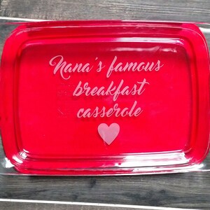 May include: A clear glass baking dish with red interior and handles. The dish has "Nana's famous breakfast casserole" etched on the front with a pink heart.