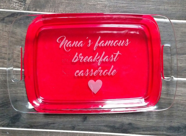 May include: A clear glass baking dish with red interior and handles. The dish has "Nana's famous breakfast casserole" etched on the front with a pink heart.