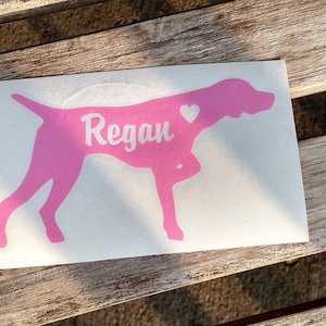 May include: Pink vinyl decal of a dog silhouette with the name "Regan" and a small white heart.