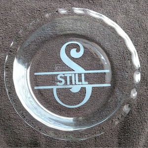 May include: A clear glass pie plate with a white etched design of the letter "S" and the word "STILL" underneath.