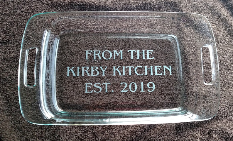 May include: A clear glass baking dish with two handles. The dish is engraved with the text "From the Kirby Kitchen Est. 2019".