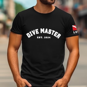 May include: Black t-shirt with white text that reads "Dive Master Est. 2024". The right sleeve has a red and white scuba diving flag.