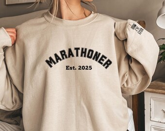 May include: A beige sweatshirt with the text "MARATHONER Est. 2025" printed on the front. The sweatshirt has a small American flag patch on the sleeve.