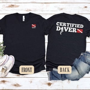 May include: Black t-shirts with scuba diving theme. One shirt has "RILEY" and a red and white dive flag on the front. The other shirt has "CERTIFIED DIVER" and a diver graphic on the back. The shirts are displayed with jeans and sneakers.