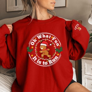 May include: Red Christmas sweatshirt with a white circle design featuring a gingerbread man running with the text "Oh What Fun It Is To Run".