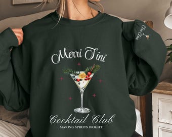 Può includere: Felpa verde con un design a tema natalizio che presenta un bicchiere da martini con bacche e un rametto di pino. Il testo "Merri Tini Cocktail Club Making Spirits Bright" è stampato sulla felpa.