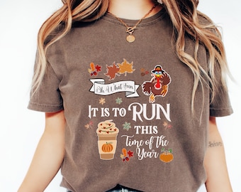May include: Brown t-shirt with a Thanksgiving-themed design. It features a turkey wearing a pilgrim hat, a coffee cup with fall-themed decorations, and the text "Oh What Fun It Is To Run This Time Of The Year".