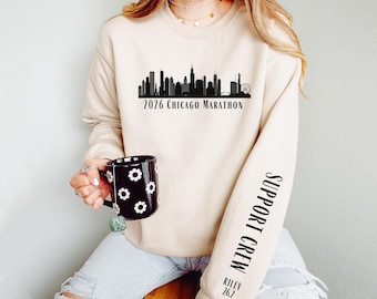 May include: A beige crewneck sweatshirt featuring a black Chicago skyline graphic with the text 2026 CHICAGO MARATHON. The left sleeve displays the text SUPPORT CREW, RILEY, and 26.2 in black lettering. The garment is paired with light blue jeans.