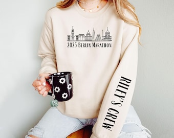 May include: A beige sweatshirt with a black graphic of the Berlin skyline and the text "2025 Berlin Marathon". The left sleeve has the text "Riley's Crew" printed vertically in black.