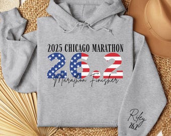 May include: A gray hooded sweatshirt with the text "2025 Chicago Marathon 26.2 Marathon Finisher" in black, blue, and red. The number 26.2 is designed with the American flag. The sleeve has the name "Riley" and "26.2" in black.