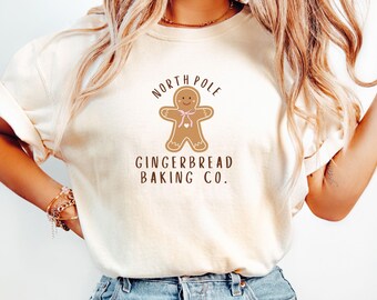 May include: Cream-colored t-shirt featuring a gingerbread man graphic with a pink bow and the text "NORTH POLE GINGERBREAD BAKING CO." in brown. The shirt is made of soft material and is suitable for casual wear.
