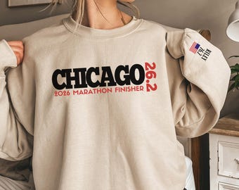 May include: A beige crewneck sweatshirt featuring the black text CHICAGO and red text 26.2 2026 MARATHON FINISHER on the chest. The left sleeve displays a small American flag icon above the text 26.2 RILEY in black.