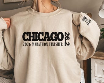 May include: A beige crewneck sweatshirt featuring black text that reads CHICAGO 26.2 2026 MARATHON FINISHER on the chest. The left sleeve includes the text RILEY 26.2 with a small flag icon. The garment has long sleeves and a ribbed neckline.