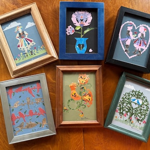Puede incluir: Una colección de seis obras de arte enmarcadas. Las obras de arte presentan varios diseños coloridos, incluyendo una flor, pájaros y figuras. Los marcos son de varios colores, incluyendo madera, plata, azul y negro. Las obras de arte están dispuestas sobre una superficie de madera.