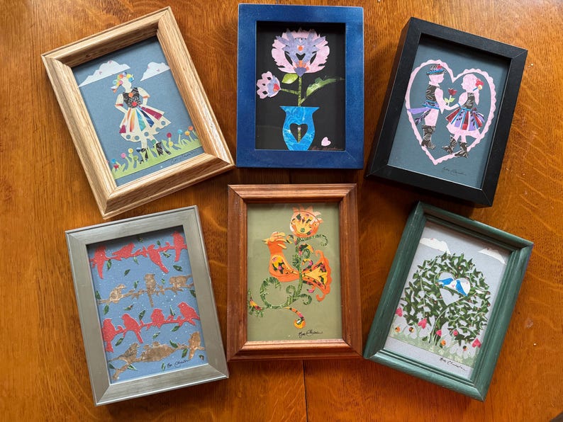 Pu&ograve; includere: Una collezione di sei opere d'arte incorniciate. Le opere presentano vari disegni colorati, tra cui un fiore, uccelli e figure. Le cornici sono di vari colori, tra cui legno, argento, blu e nero. Le opere d'arte sono disposte su una superficie di legno.