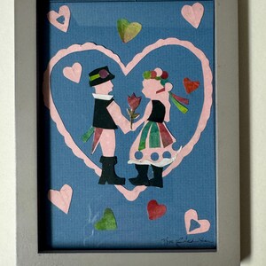 Pu&ograve; includere: Un'opera d'arte popolare incorniciata raffigurante una coppia che si tiene per mano all'interno di un cuore rosa su sfondo blu. La coppia indossa abiti tradizionali, con un fiore tra loro. Cuori rosa, verdi e rossi circondano l'immagine centrale.
