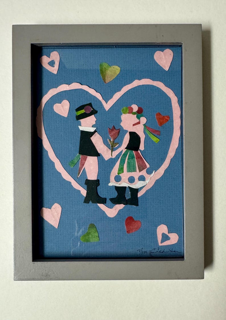 Pu&ograve; includere: Un'opera d'arte popolare incorniciata raffigurante una coppia che si tiene per mano all'interno di un cuore rosa su sfondo blu. La coppia indossa abiti tradizionali, con un fiore tra loro. Cuori rosa, verdi e rossi circondano l'immagine centrale.