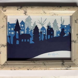 Pu&ograve; includere: Un'opera d'arte incorniciata con una silhouette di una citt&agrave; con case e alberi in varie tonalit&agrave; di blu. L'opera &egrave; ambientata su uno sfondo bianco e racchiusa in una cornice beige con un motivo marmorizzato.
