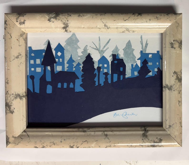 Pu&ograve; includere: Un'opera d'arte incorniciata con una silhouette di una citt&agrave; con case e alberi in varie tonalit&agrave; di blu. L'opera &egrave; ambientata su uno sfondo bianco e racchiusa in una cornice beige con un motivo marmorizzato.