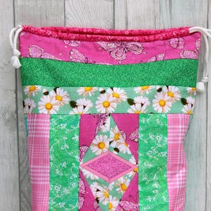 May include: A pink and green drawstring bag with a patchwork design featuring daisies, butterflies, and a diamond shape. The bag is made of fabric and has a white drawstring.