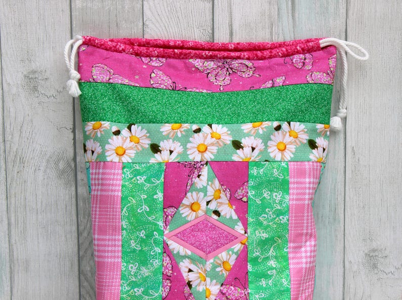May include: A pink and green drawstring bag with a patchwork design featuring daisies, butterflies, and a diamond shape. The bag is made of fabric and has a white drawstring.