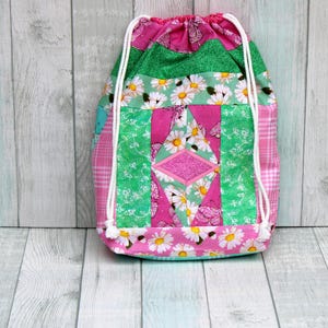 May include: A pink and green drawstring bag with a floral pattern and a diamond-shaped patch. The bag is made of fabric and has a white drawstring.