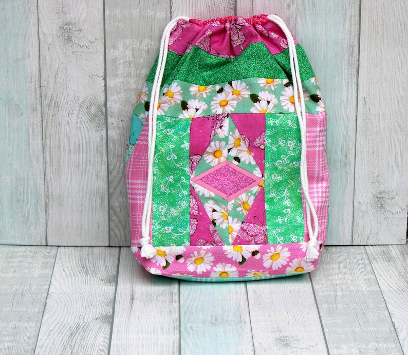 May include: A pink and green drawstring bag with a floral pattern and a diamond-shaped patch. The bag is made of fabric and has a white drawstring.