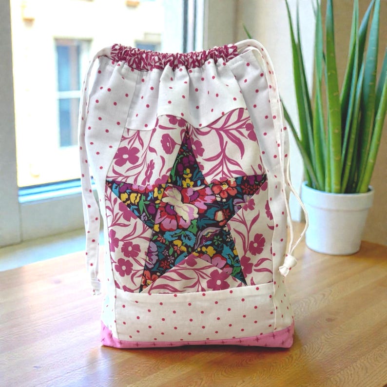 Pode incluir: Uma bolsa com cord&atilde;o com um design de estrela de patchwork. A bolsa apresenta um fundo branco com bolinhas rosa e detalhes florais. A estrela &eacute; preenchida com um padr&atilde;o floral colorido. A parte inferior e o cord&atilde;o s&atilde;o rosa.