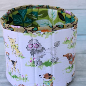 May include: A white fabric storage bin with a green and brown floral print on the inside. The outside of the bin features a white background with a repeating pattern of dogs wearing glasses and hats.