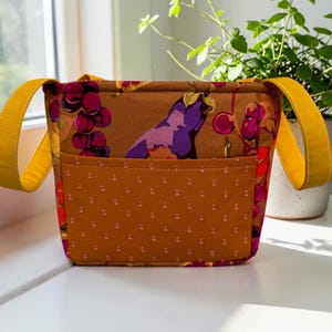 May include: A fabric tote bag featuring a mustard yellow exterior pocket with a subtle dotted texture. The main body of the bag displays a vibrant print of purple grapes and eggplants on a brown background, complemented by two mustard yellow straps.