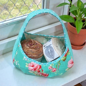 May include: A quilted fabric basket with a handle, featuring a floral pattern of pink roses on a turquoise background. The basket contains two spools of yarn, one brown and one white.