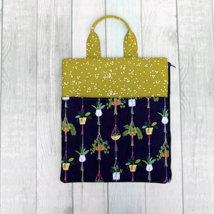 May include: A fabric tote bag with a green handle and top section, and a navy blue bottom section. The top section has a pattern of small white dots. The bottom section features a pattern of hanging potted plants.