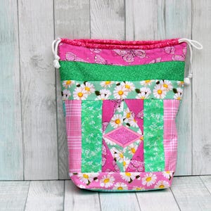 May include: A pink and green drawstring bag with a patchwork design featuring daisies, butterflies, and a diamond shape. The bag is made of fabric and has a white drawstring.