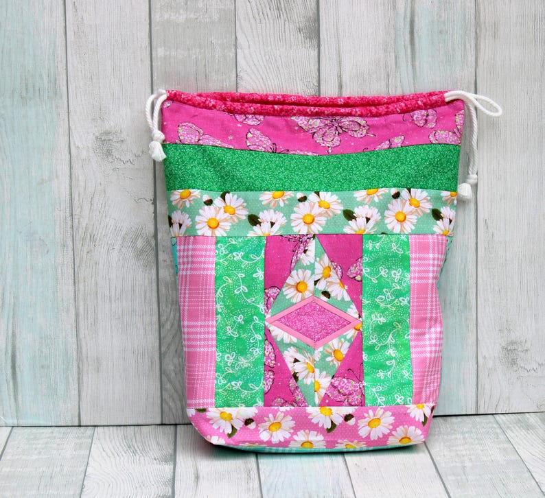 May include: A pink and green drawstring bag with a patchwork design featuring daisies, butterflies, and a diamond shape. The bag is made of fabric and has a white drawstring.