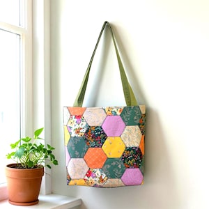 May include: A handmade tote bag with a patchwork design of colorful hexagonal fabric pieces. The bag features a variety of floral patterns in shades of pink, yellow, orange, and green. It has olive green straps and is hanging against a white wall.